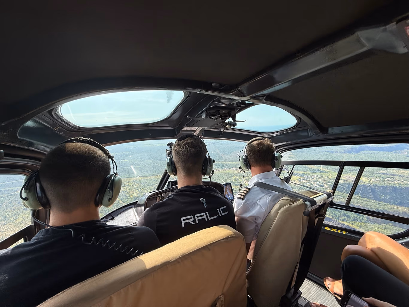 Three founders in aviation headsets seated behind the pilot in a helicopter, looking out over the landscape far below — someone else flying