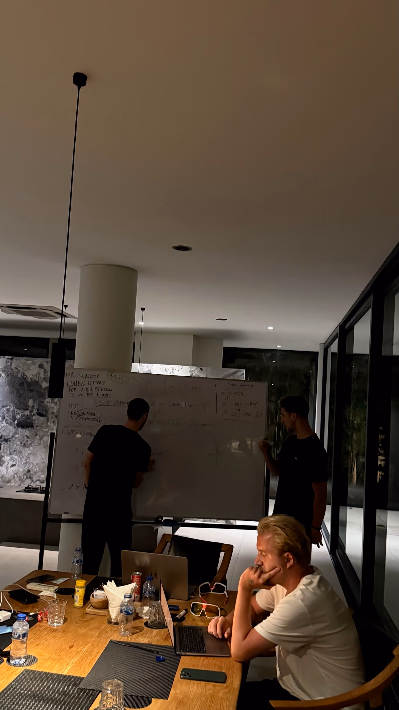 Three founders working late in a villa at night — two at a whiteboard full of notes, one at a table on a laptop, glass walls behind
