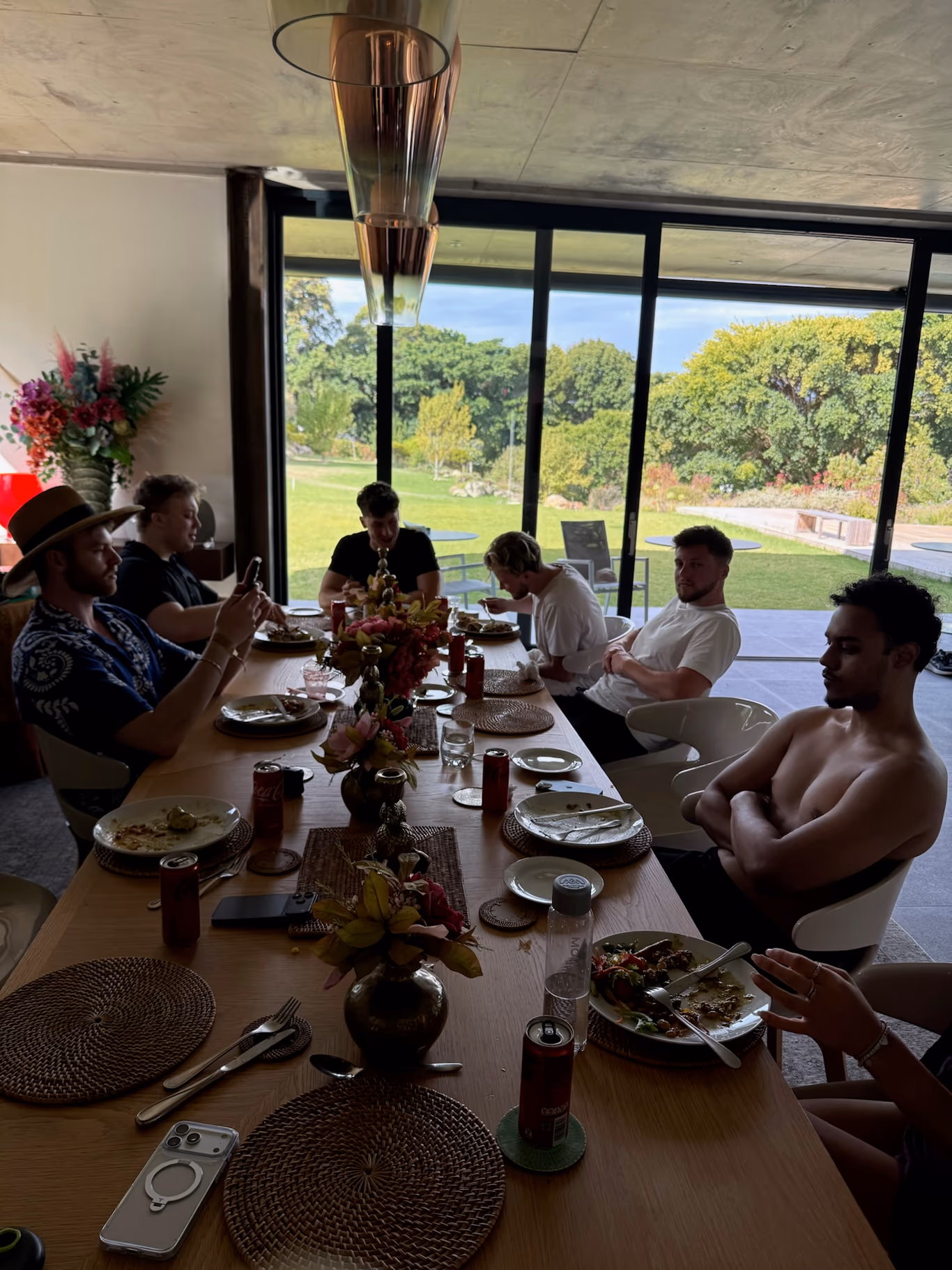 Founder support system — the table that has your back Founders gathered around a long wooden table for a midday meal, floor-to-ceiling windows behind them framing a green garden