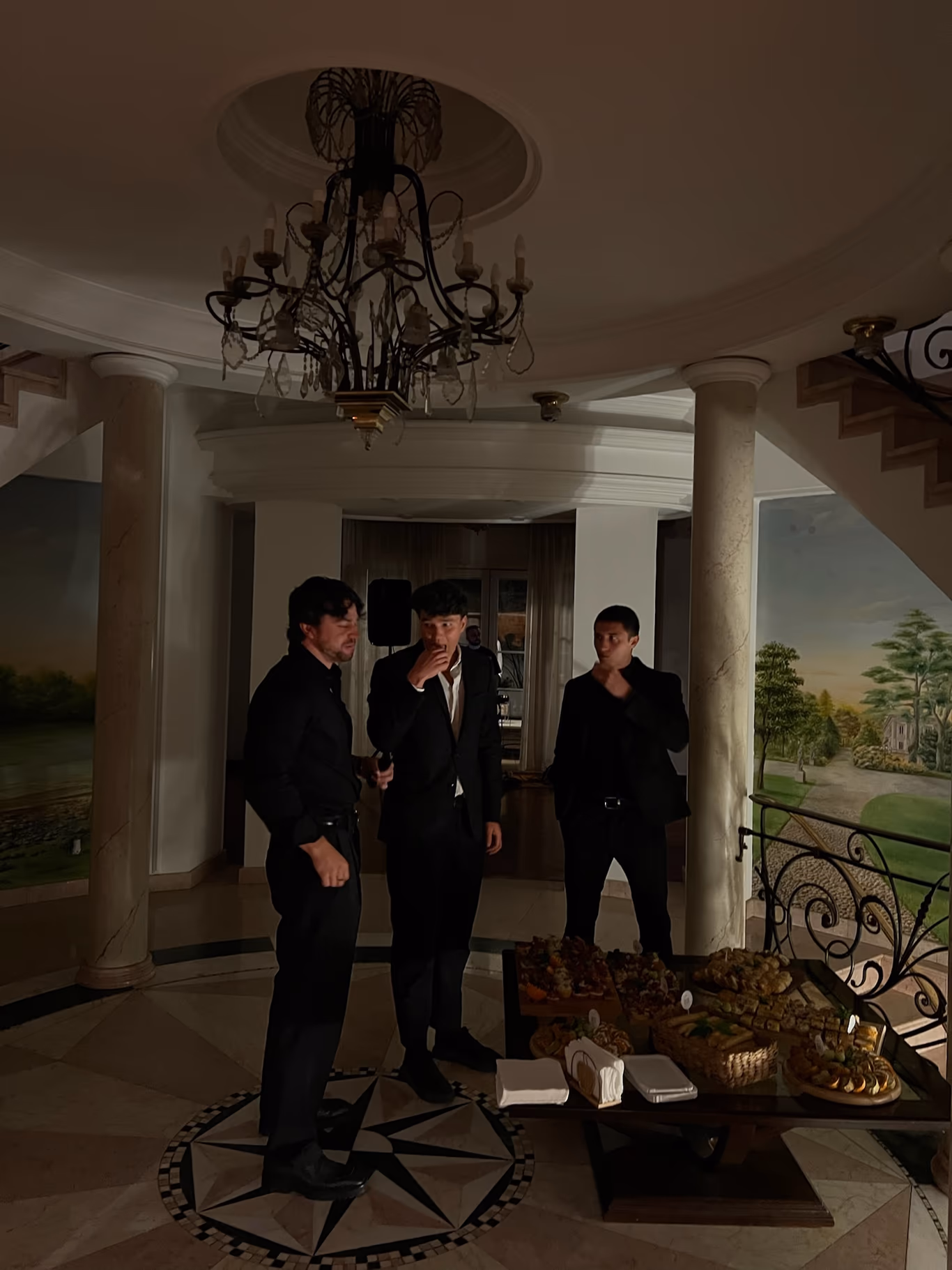 Three founders in dark suits standing and talking around a food-laden table under a chandelier in a villa interior