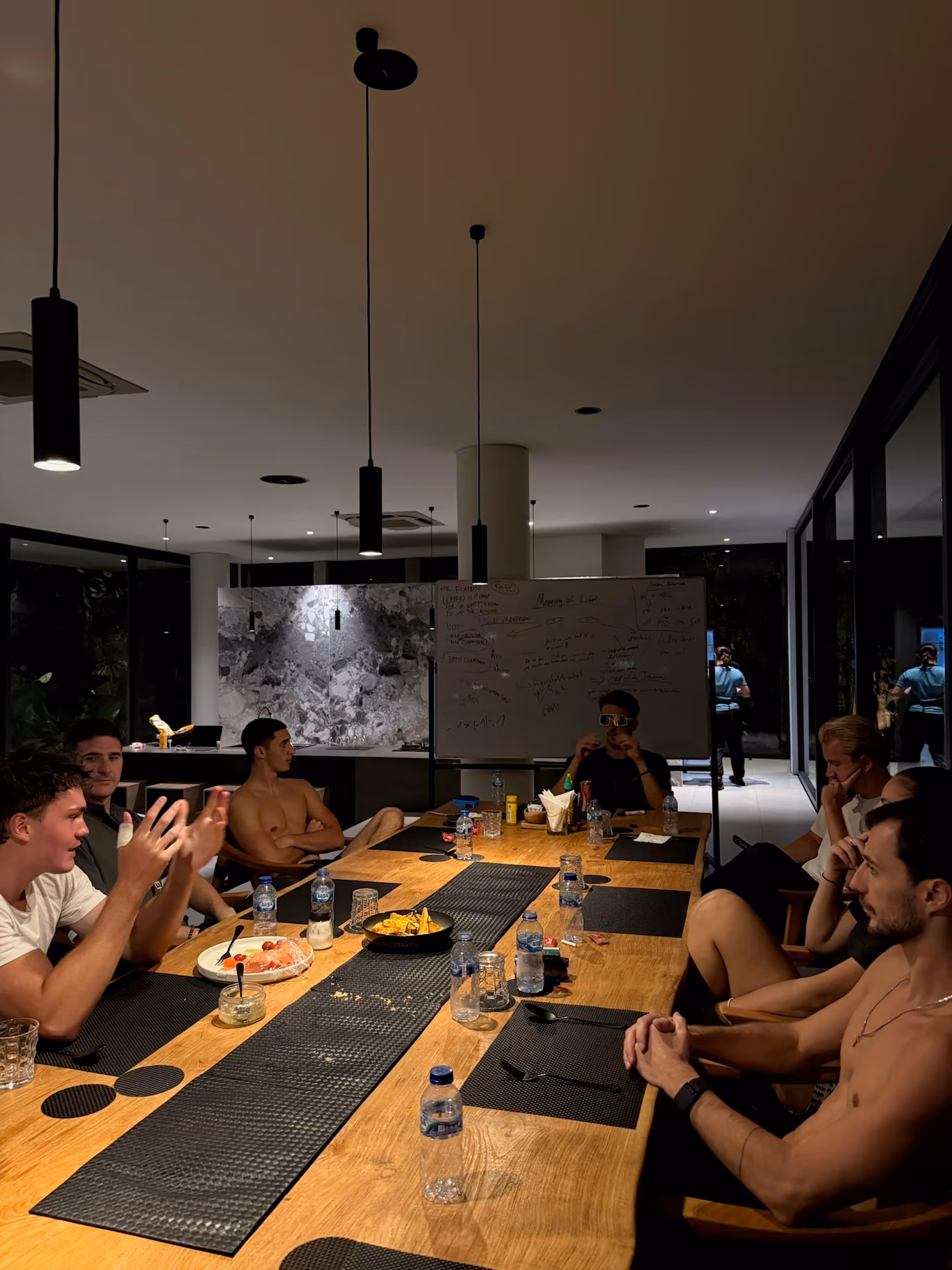 Join Helix — the welcome after the vetting Founders seated around a long table late at night at a Helix villa, one mid-gesture, a whiteboard of notes behind them